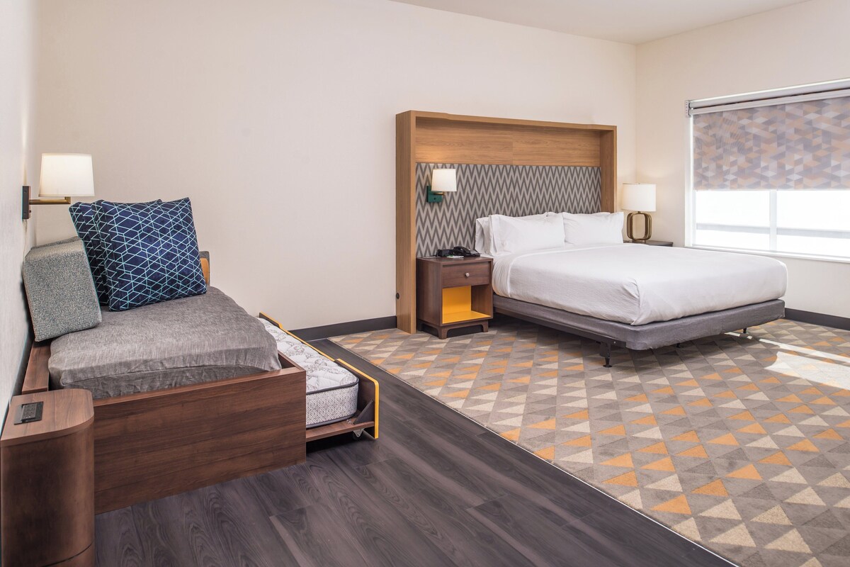 A hotel room with a bed, a sofa bed, a nightstand, a lamp, and a patterned floor.