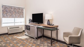 Ein Hotelzimmer mit Schreibtisch, Stuhl, Fernseher, Lampe, Klimaanlage und einem gemusterten Teppich.