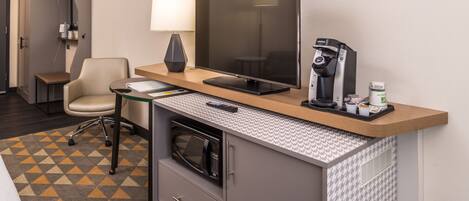 A hotel room with a desk, a chair, a flat-screen TV, a coffee maker, a microwave, and a patterned rug.