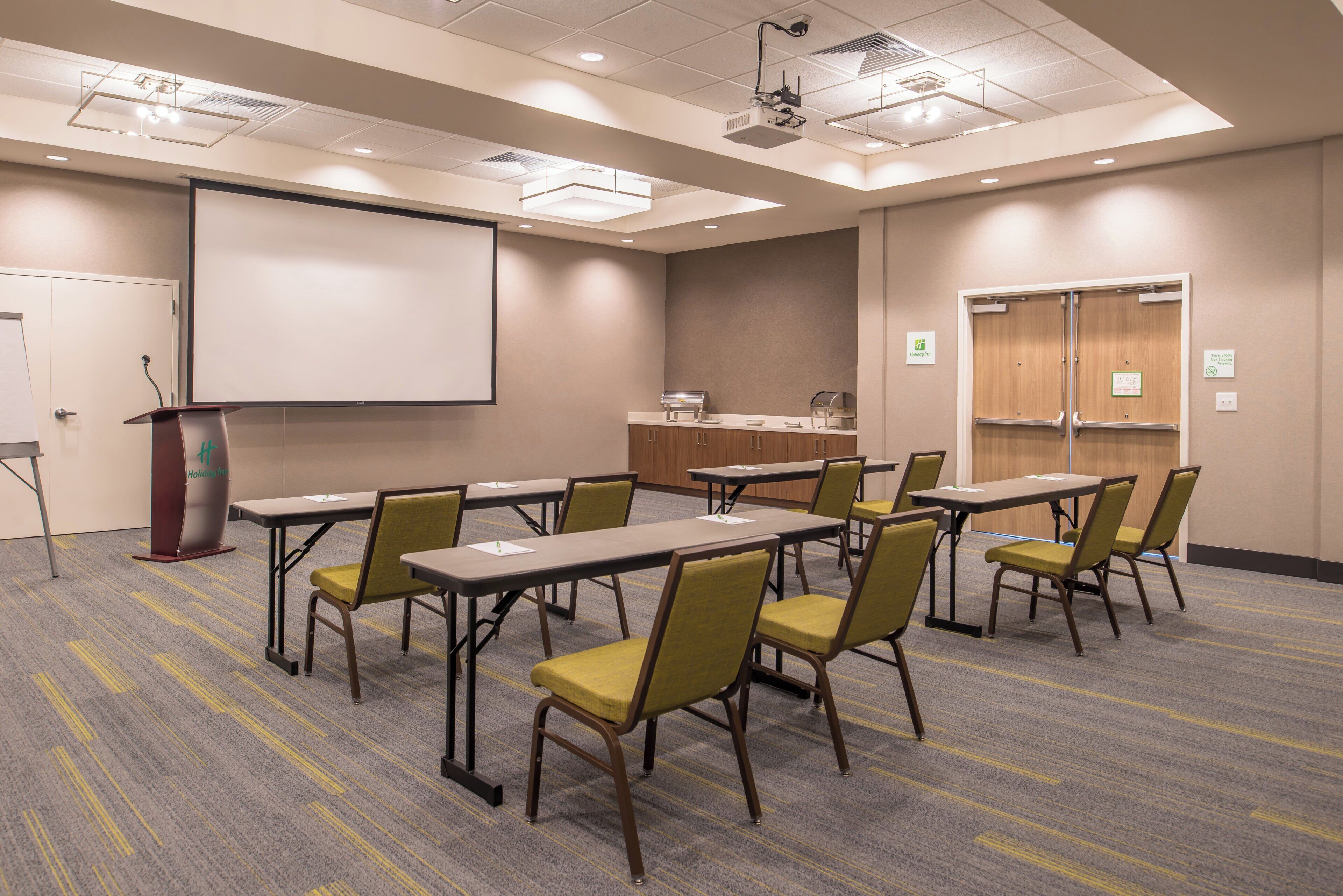 Un salón de conferencias con pantalla de proyector, mesas, sillas y un atril.