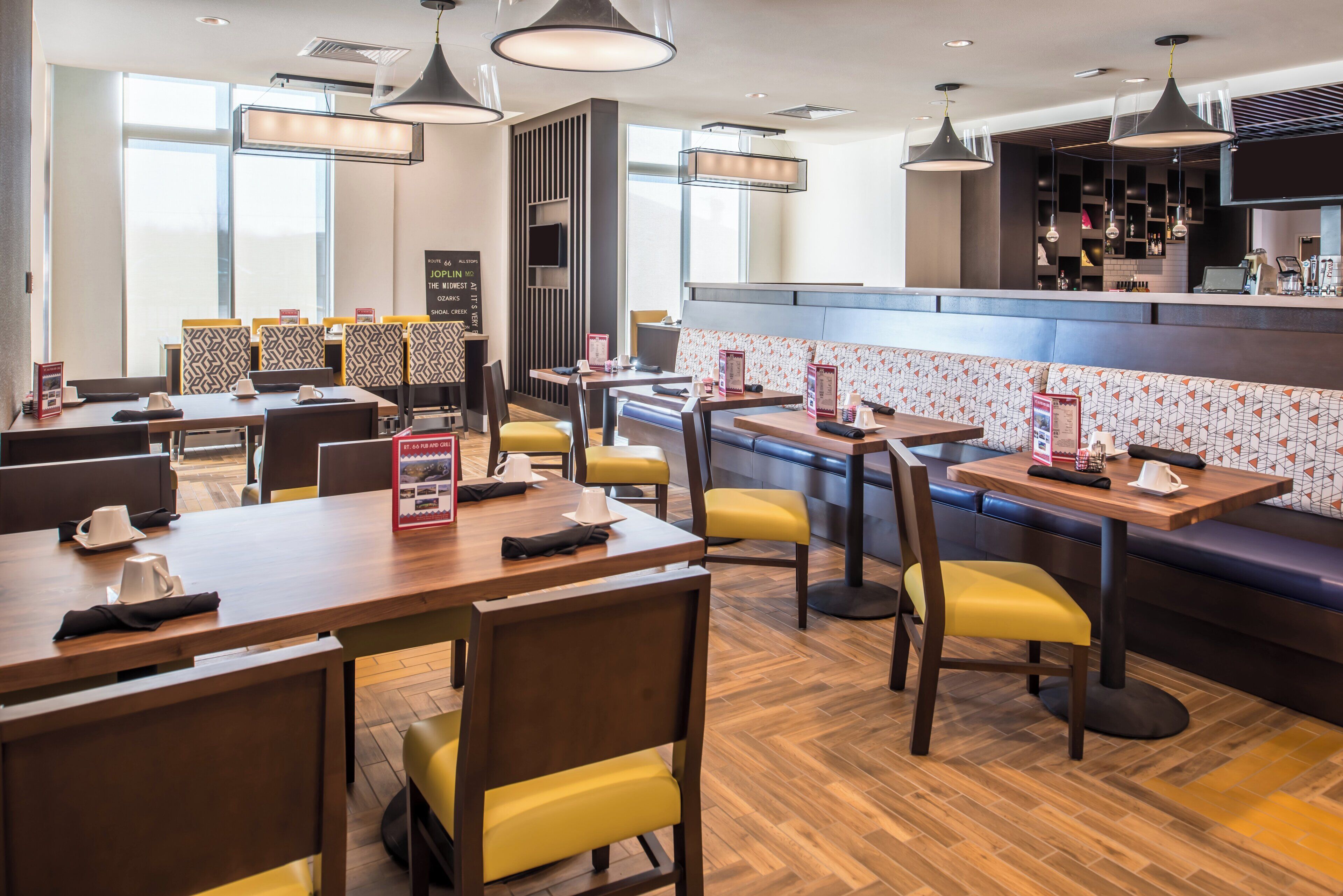 A modern restaurant with wooden tables and yellow chairs, a bar area, and a menu board.