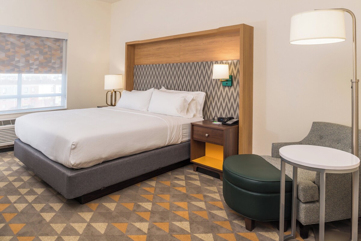 Une chambre d’hôtel avec un lit, une table de chevet, une lampe, une chaise et un tapis à motifs.