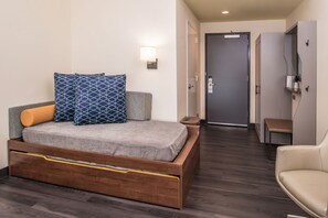 Une chambre d’hôtel moderne avec un lit en bois, un banc, une lampe fixée au mur et une porte donnant accès à une salle de bain.