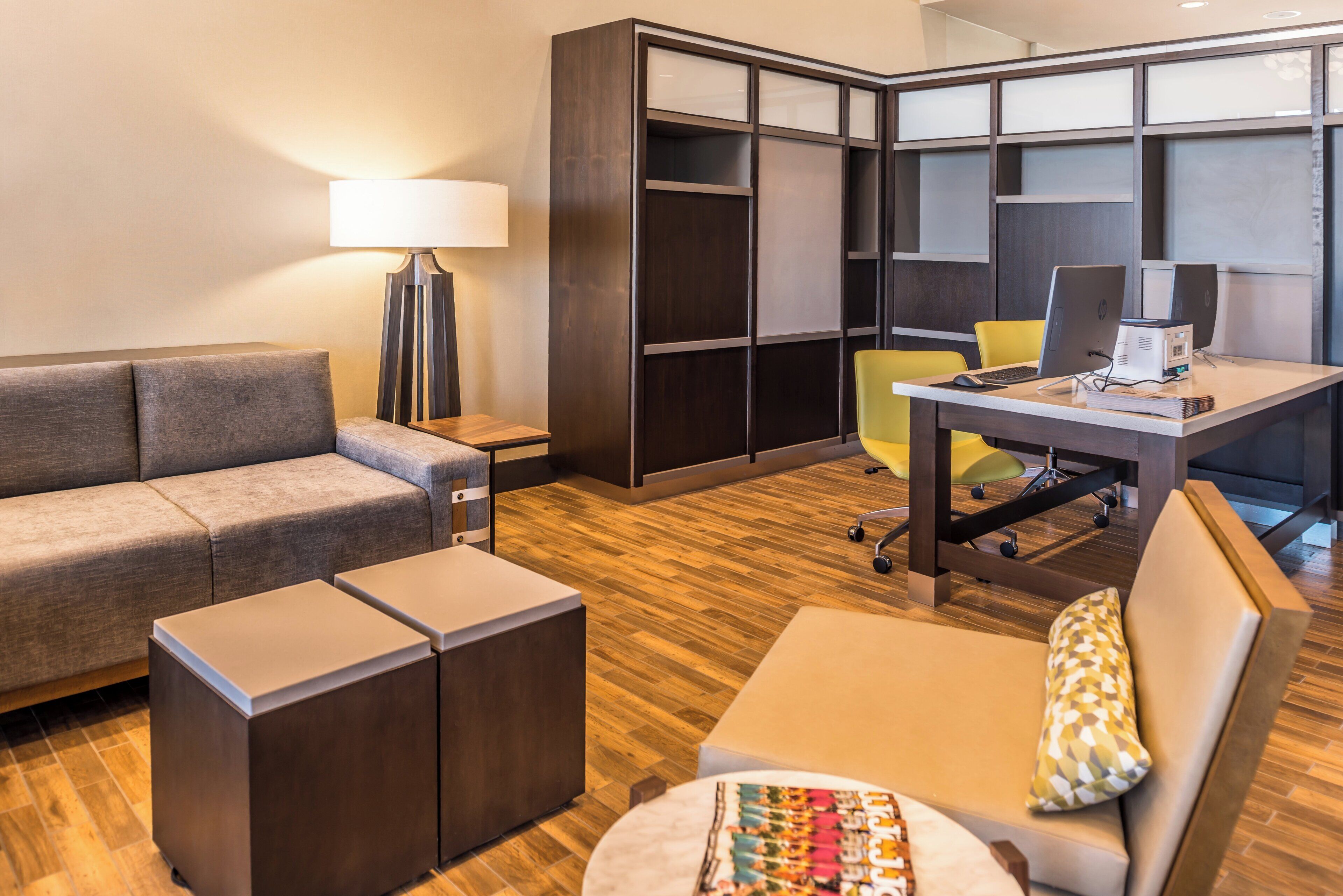 A modern hotel room with a sofa, a desk with a computer, a lamp, and a storage unit.