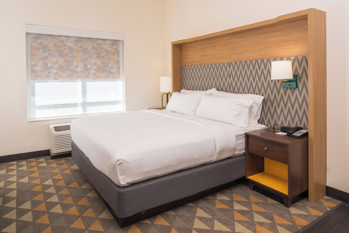 Une chambre d’hôtel avec un lit, une table de chevet, une lampe, une fenêtre avec des stores et un sol à motifs.