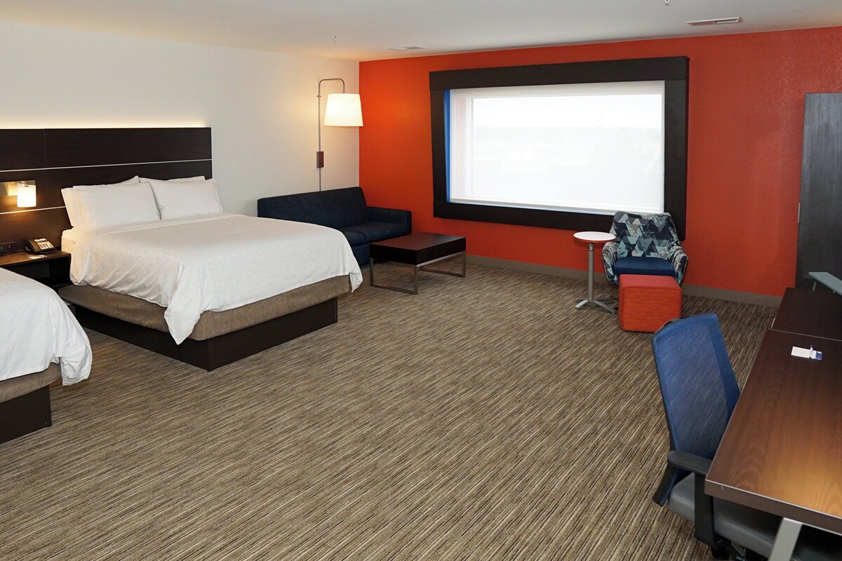A hotel room with a large bed, a desk, a chair, a small table, a sofa, a lamp, and a projector screen.