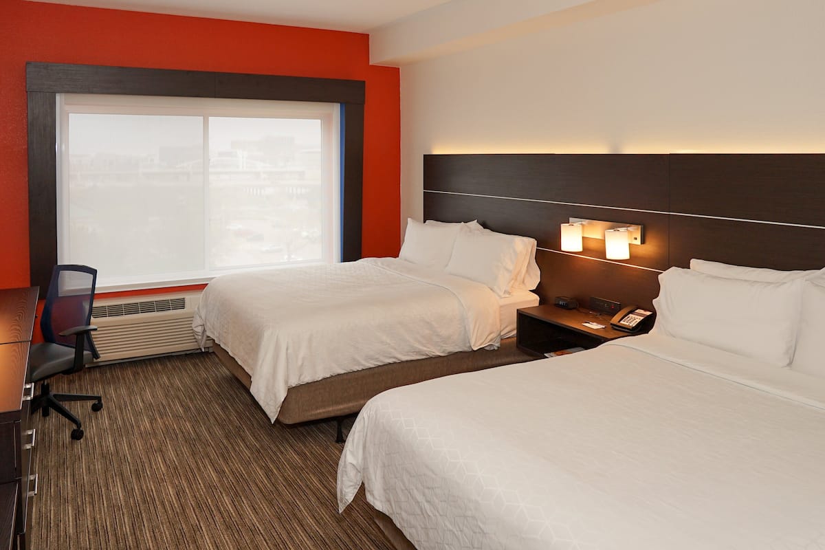 A hotel room with two beds, a desk, and a chair. There is a window with a view of buildings.