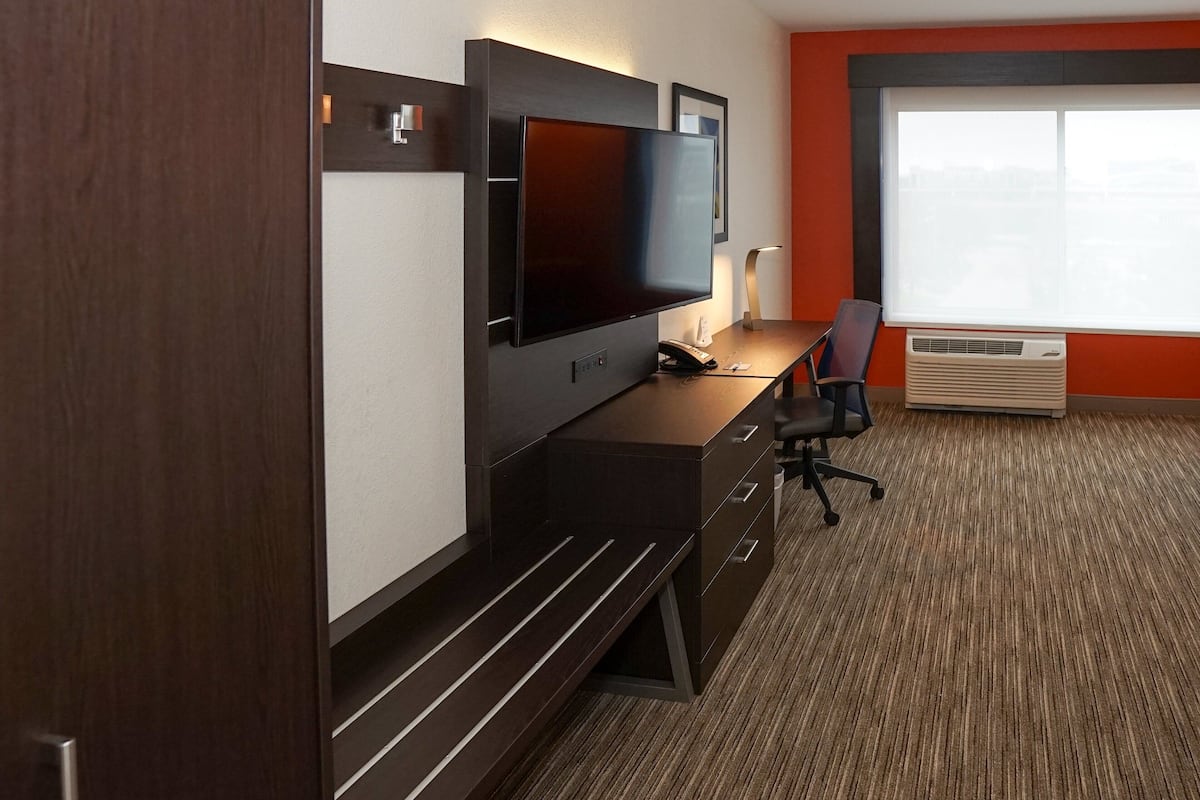 A modern hotel room with a flat-screen TV, a desk with a chair, a bench, and a large window with an air conditioning unit.