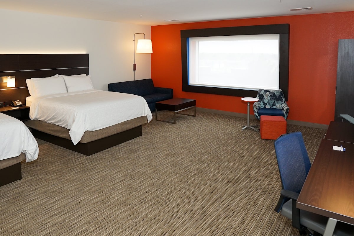 A hotel room with a large bed, a desk, a chair, a small table, a sofa, a lamp, and a projector screen.
