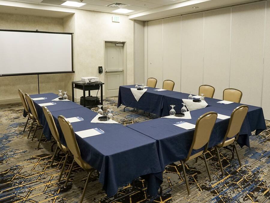 Una sala de conferencias con una gran mesa, sillas, una pantalla de proyector y piso alfombrado.