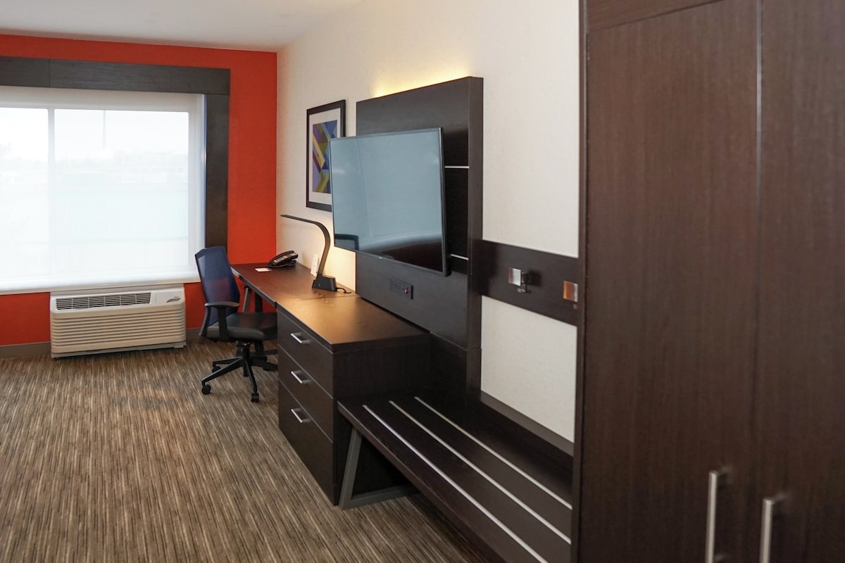 A modern hotel room with a large window, a desk with a computer, a chair, and a wardrobe.