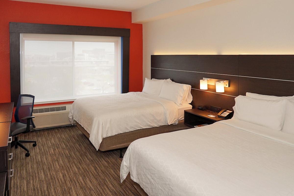A hotel room with two beds, a desk, and a chair. There is a window with a view of buildings.