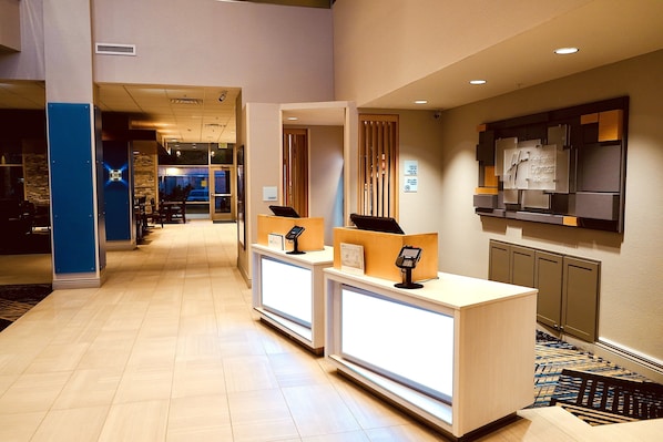 A hotel reception area with two reception desks, a digital check-in kiosk, and a wall-mounted information board.