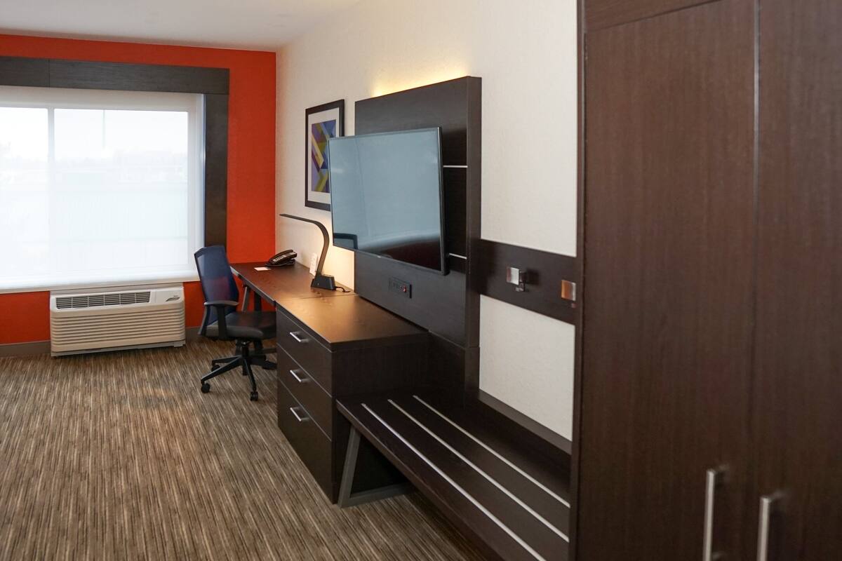 A modern hotel room with a large window, a desk with a computer, a chair, and a wardrobe.