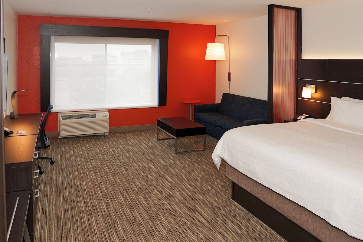 A hotel room with a large bed, a sofa, a small table, a desk with a chair, a window with blinds, and a wall-mounted lamp.