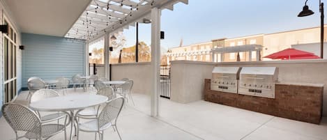Een overdekt terras met een witte tafel en stoelen, een bakstenen muur met barbecues, en een pergola met lichtslingers.