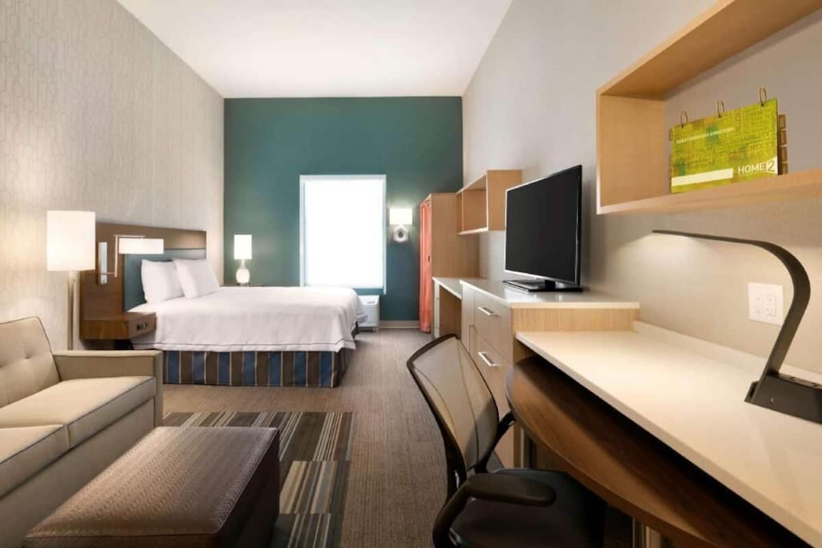 A hotel room with a bed, desk, chair, TV, and a bookshelf.