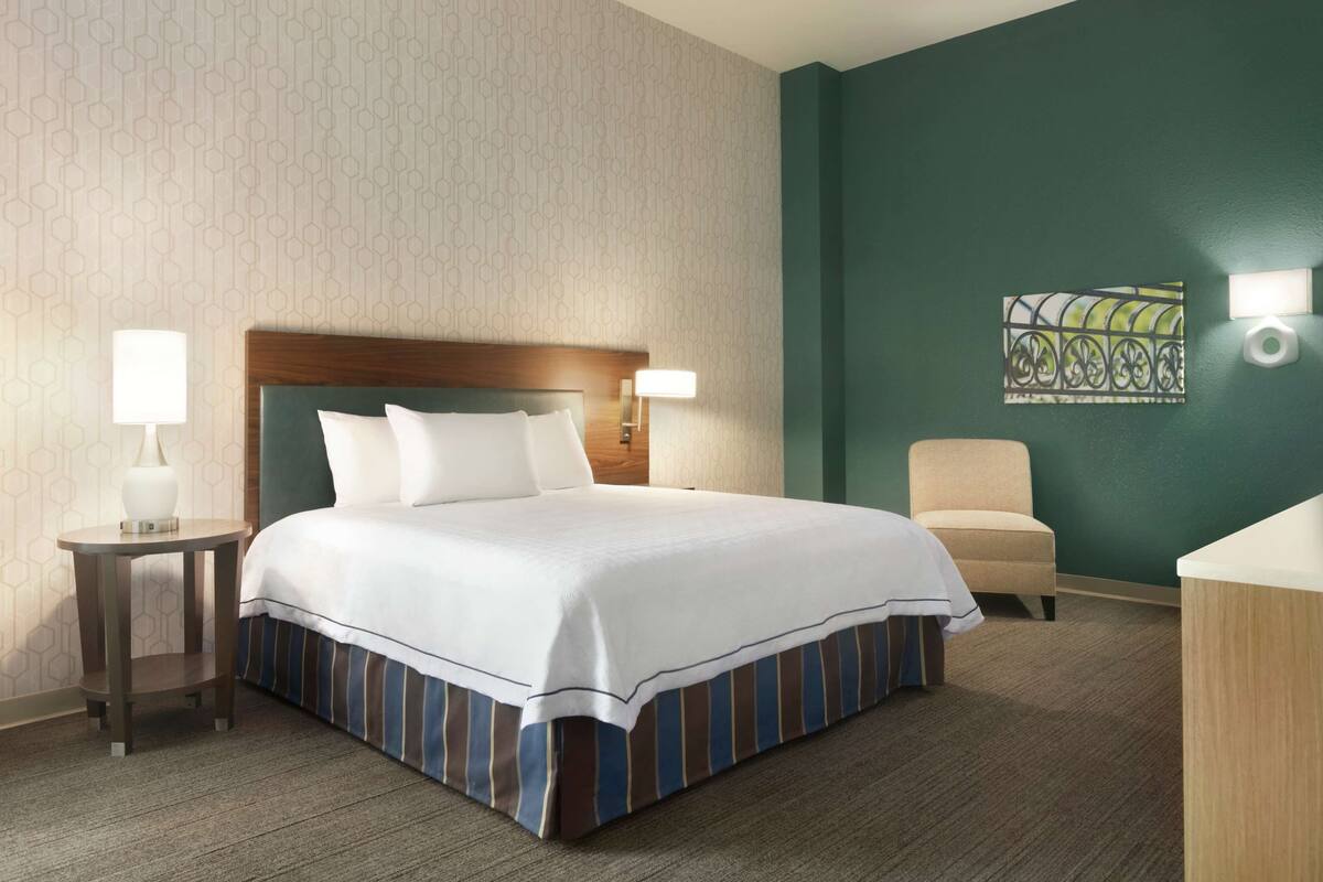 A hotel room with a large bed, two bedside lamps, a chair, a small table, and a decorative wall piece.
