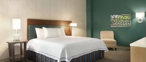 Habitación de hotel con una cama grande, dos lámparas de noche, una silla, una mesita y un adorno en la pared.