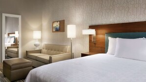 Ein Hotelzimmer mit einem großen Bett, einer Couch, einem Sessel und einem kleinen Tisch. Zwei Wandlampen und ein Gemälde sind an der Wand angebracht.