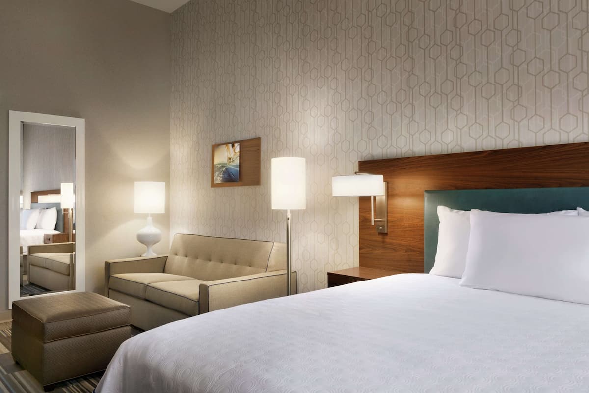 A hotel room with a large bed, a sofa, a chair, and a small table. There are two wall-mounted lamps and a painting on the wall.