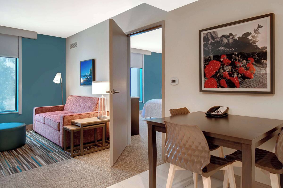 A hotel room with a sofa, a small table, a lamp, a dining table with chairs, and a framed picture on the wall.