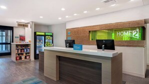Zona de recepción de hotel con diseño moderno, que incluye un mostrador de recepción, una pantalla digital mostrando el nombre del hotel, un refrigerador con bebidas y un quiosco de autoservicio.