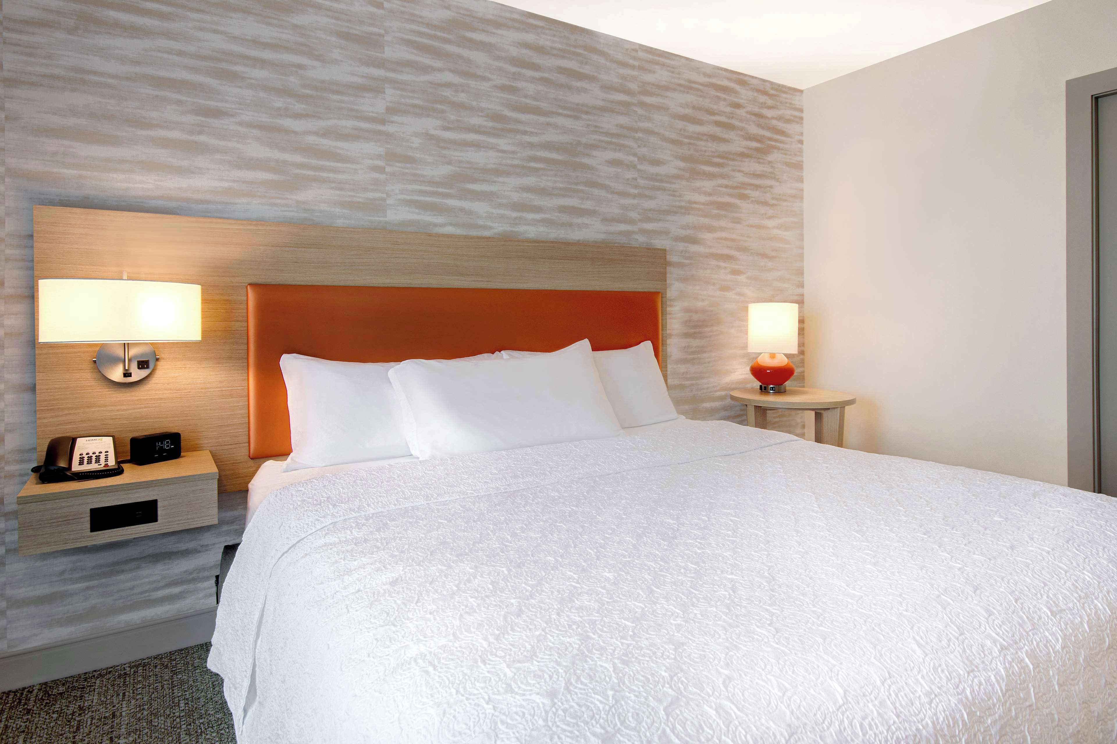 Une chambre d’hôtel avec un grand lit, deux tables de chevet avec des lampes, une petite table de nuit avec un téléphone et une horloge, et un mur à texture.