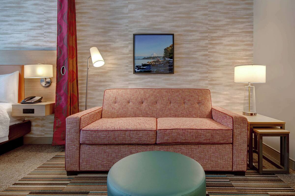 A hotel room with a bed, a sofa, a round ottoman, two bedside tables with lamps, a phone, and a framed picture on the wall.