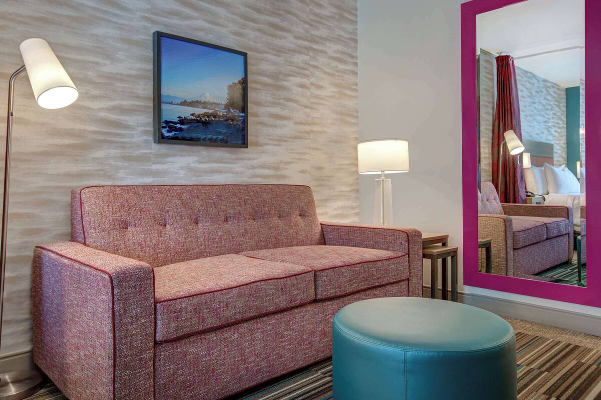 A hotel room with a red sofa, a blue ottoman, a floor lamp, a table lamp, and a large mirror reflecting the room.