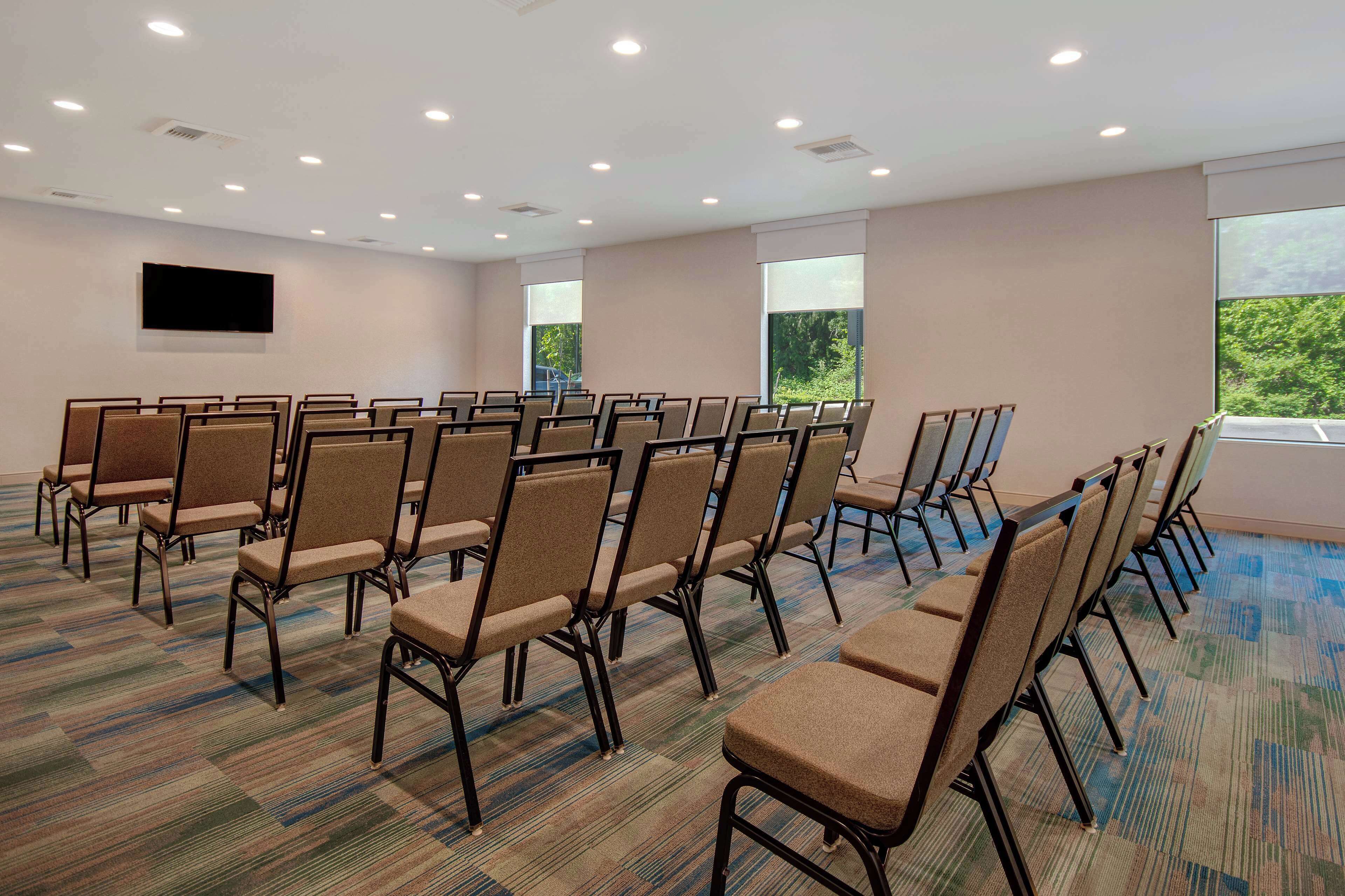 A conference room with rows of chairs, a flat-screen TV on the wall, and large windows with a view of greenery.