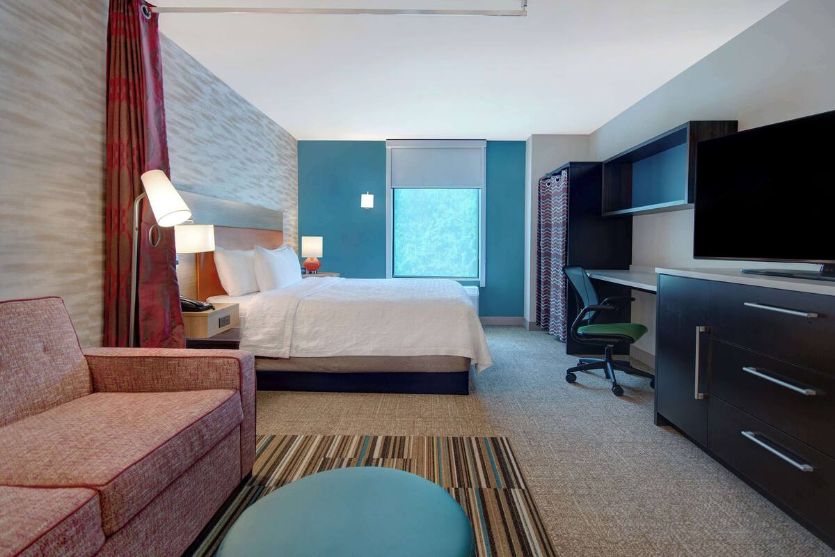 A hotel room with a bed, a sofa, a desk with a TV, and a window.