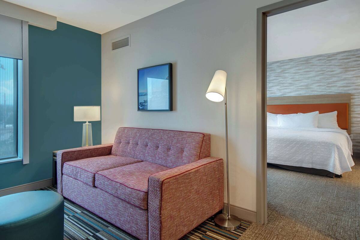 A hotel room with a pink sofa, a standing lamp, a framed picture on the wall, and a bed with white bedding.
