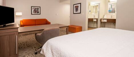 A hotel room with a bed, a desk, a chair, a television, and a bathroom with a mirror and sink.