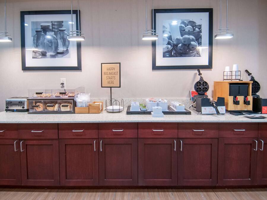 Una barra de desayuno con diferentes alimentos y electrodomésticos, fotografías enmarcadas en la pared y un letrero que indica el servicio de desayuno.