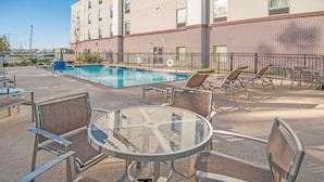 A poolside area with lounge chairs, a glass table, and a brick building in the background.