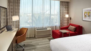 A hotel room with a bed, a desk with a laptop, a red armchair, a red sofa, and a large window with curtains.