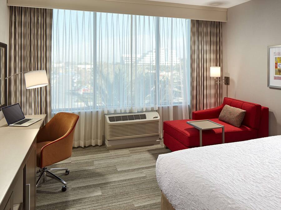 A hotel room with a bed, a desk with a laptop, a red armchair, a red sofa, and a large window with curtains.