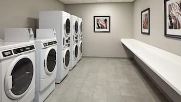 A laundry room with multiple washing machines and dryers, a white bench, and framed artwork on the wall.