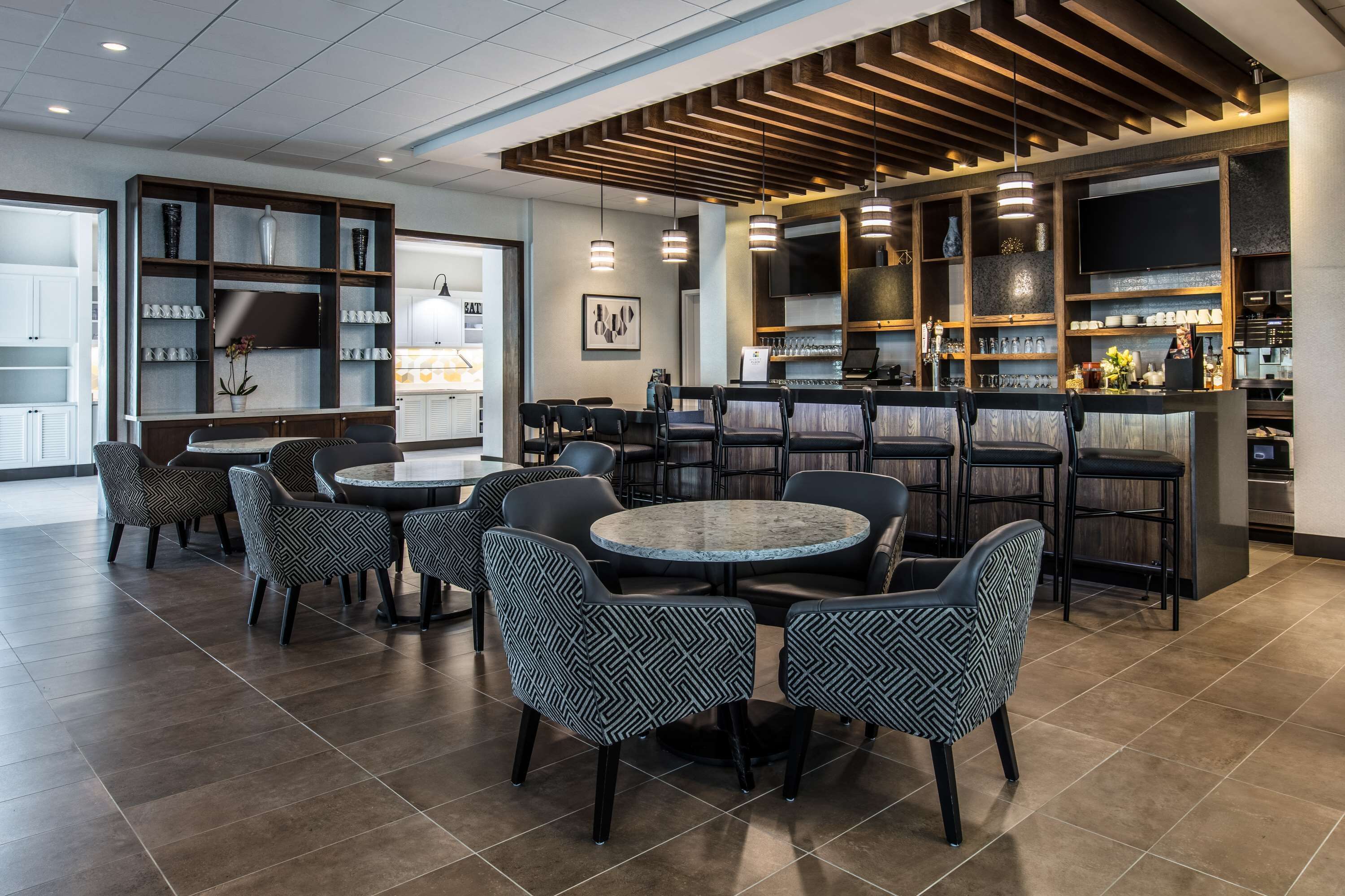 A modern bar area with round tables and patterned chairs, a wooden ceiling, and shelves with glassware and decorative items.