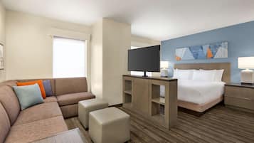 A hotel room with a sofa, ottoman, TV, bed, nightstand, lamp, and artwork on the wall.
