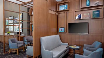 Una habitación de hotel moderna con un sofá gris, dos sillones, una pequeña mesa y una estantería con cuadros enmarcados.