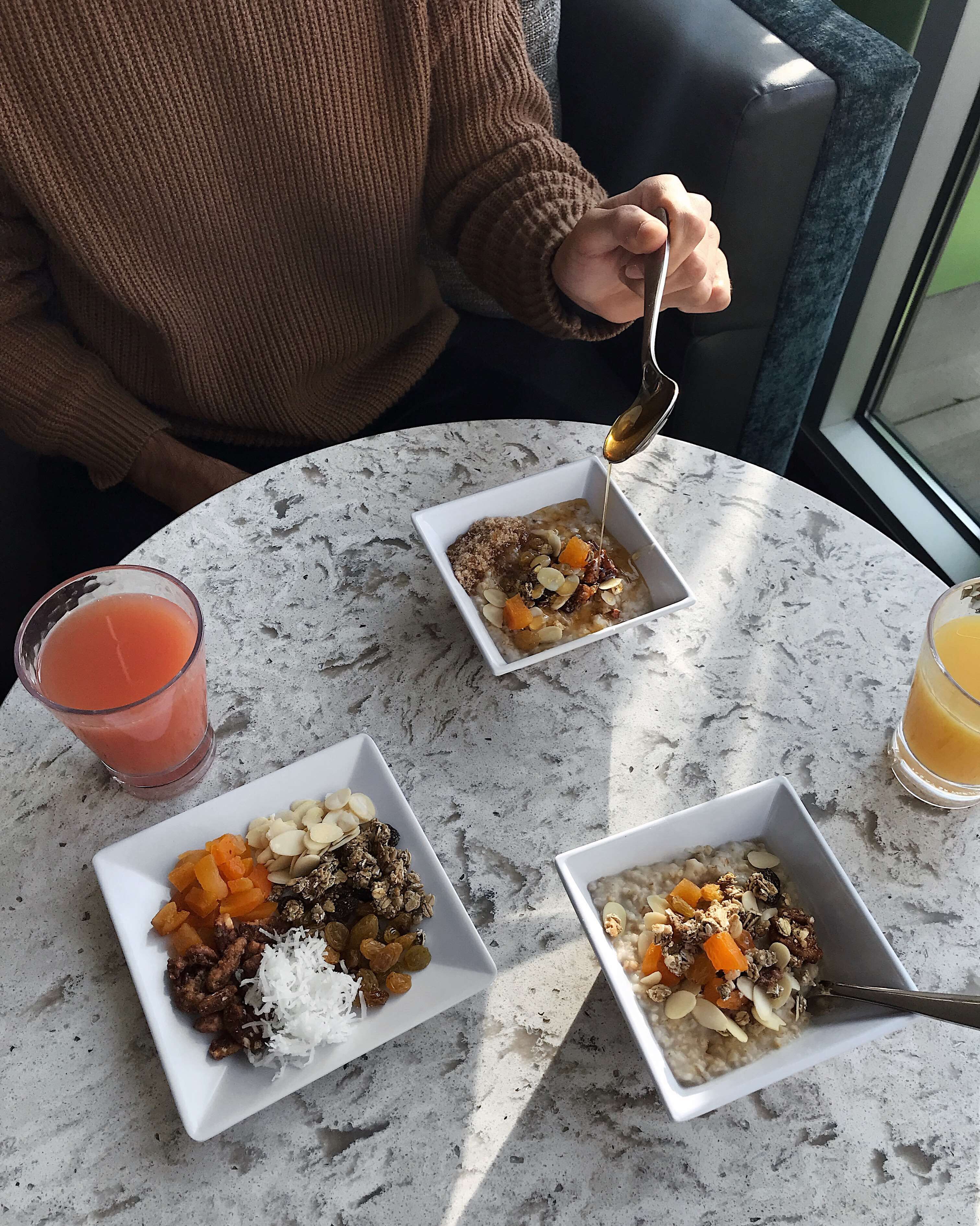 A table with three bowls of cereal topped with fruits and nuts, a glass of orange juice, and a person holding a spoon.