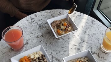 A table with three bowls of cereal topped with fruits and nuts, a glass of orange juice, and a person holding a spoon.