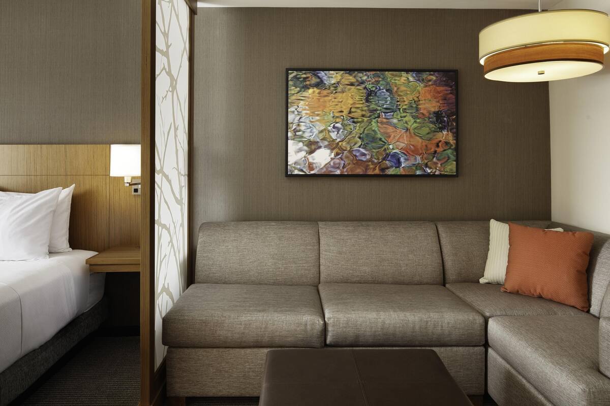 A hotel room with a sofa, a small table, a bed with white bedding, a wall-mounted painting, and a ceiling light fixture.