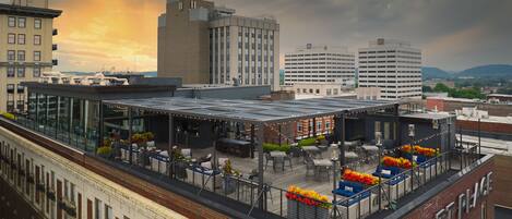 A rooftop bar with outdoor seating, a glass enclosure, and a view of the city skyline.