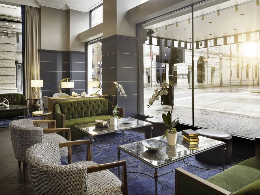 A modern lobby with a green sofa, wicker chairs, a glass coffee table, and a large window offering a view of the street.