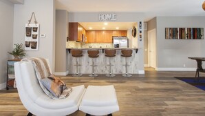 Una sala moderna con un sofá y reposapiés blancos, una cocina al fondo y un letrero con la palabra 'HOME'.