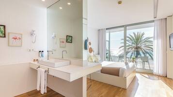 A modern hotel room with a large mirror, a white vanity, a bed with a view of the balcony, and a large window.