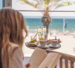 Una persona leyendo un libro en una mesa con una bandeja de comida y bebidas, con vista a una playa.